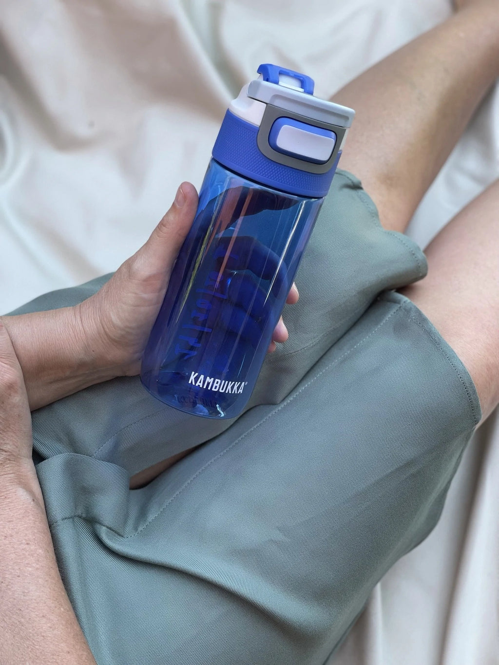Mão a segurar a Garrafa Kambukka Elton 500ml azul transparente cheia de água, com as pernas de uma pessoa em fundo.
