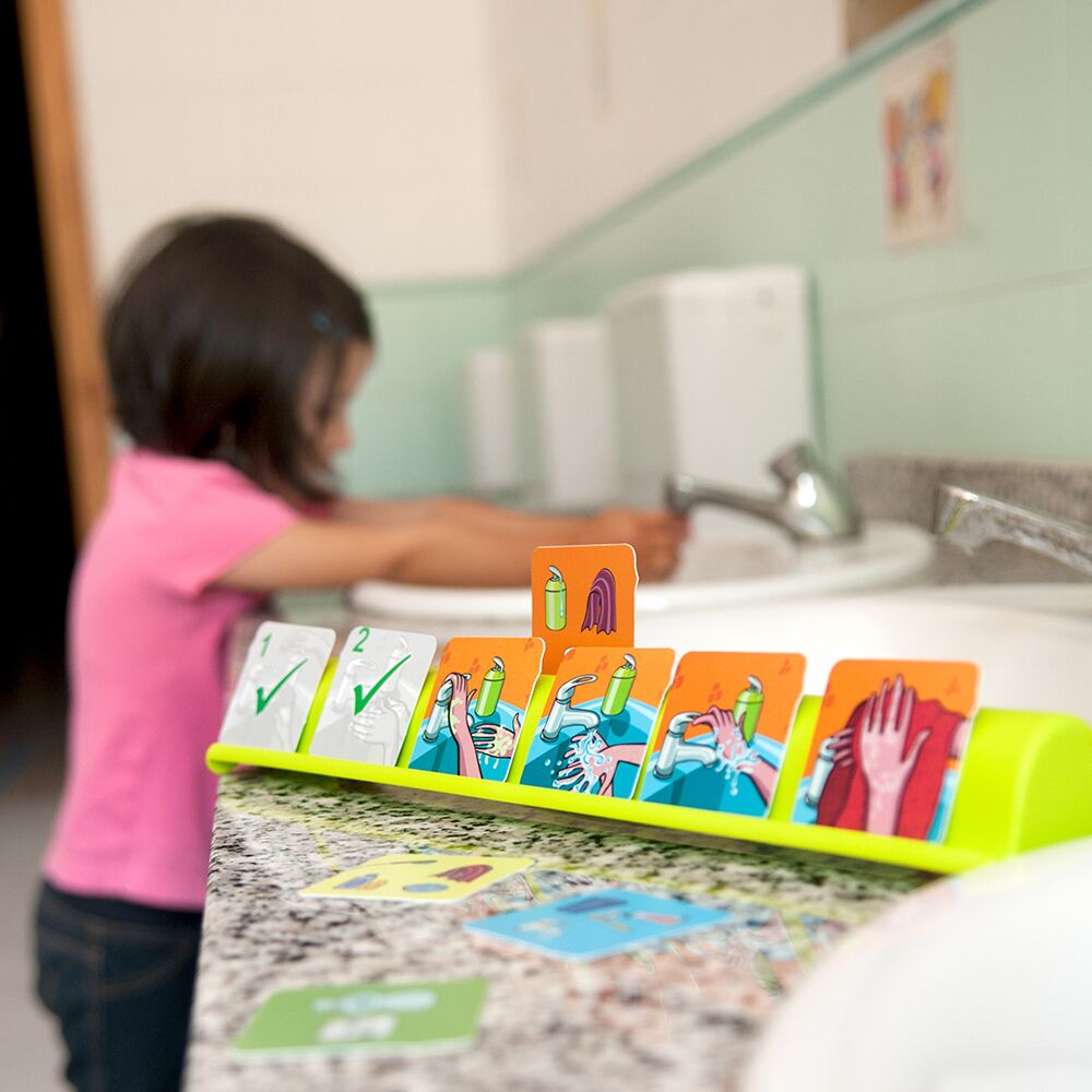 Criança a lavar as mãos na casa de banho, enquanto um jogo Miniland de sequências de hábitos de higiene está no balcão, mostrando cartões de lavar as mãos.