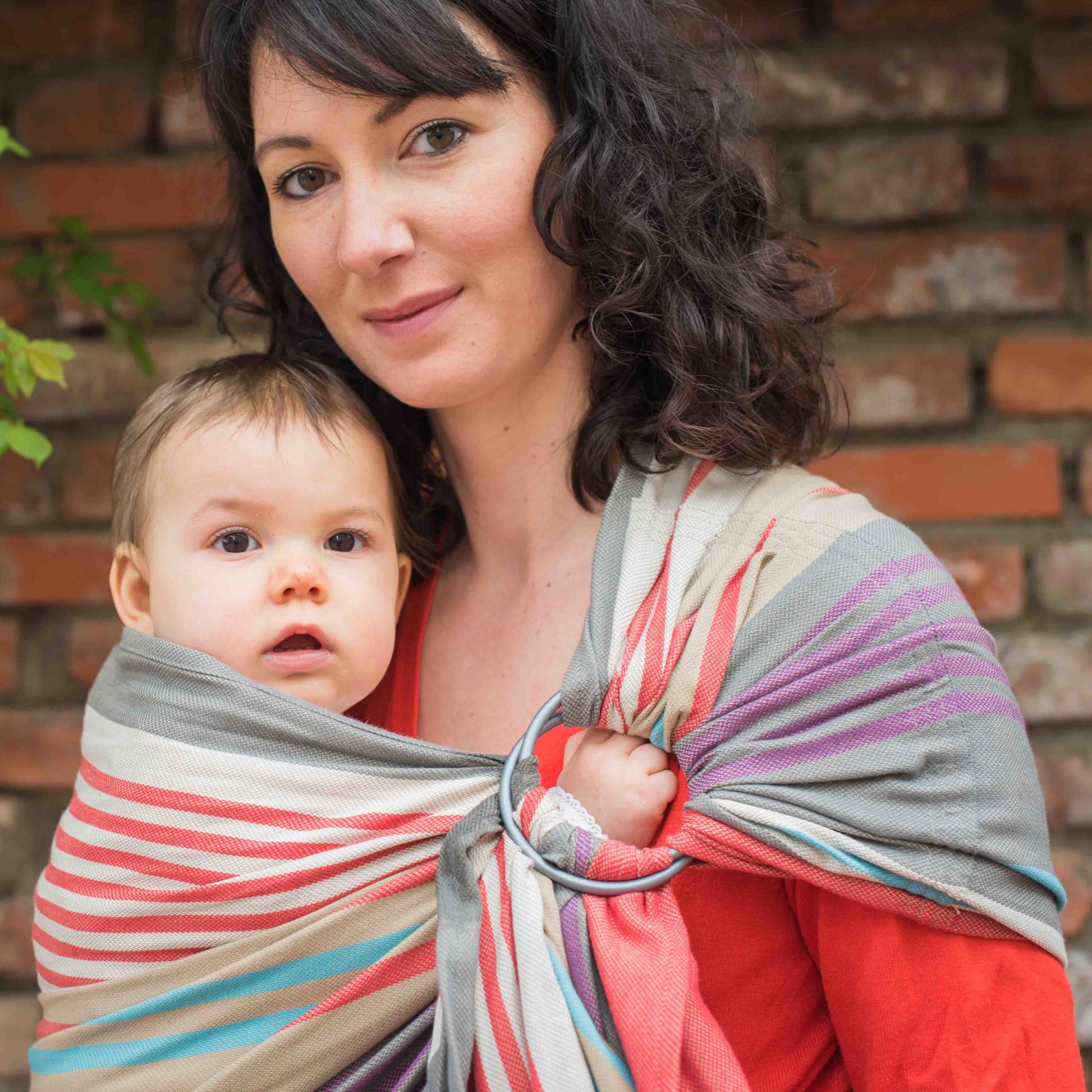 Mulher a usar um sling de argolas Neobulle JACQUARD Zèbre Beige, transportando um bebé. O sling tem um padrão colorido de listras em tons de cinza, vermelho e azul. A mulher está vestida com uma camisola vermelha e o bebé tem uma expressão curiosa. Fundo de tijolos à vista.
