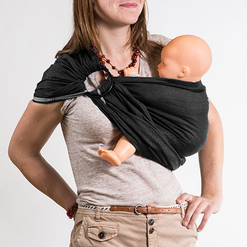 Mulher a usar um sling de argolas preto da Mamagoló, feito de linho, com um boneco a simular um bebé no interior, demonstrando a forma correta de uso do sling.