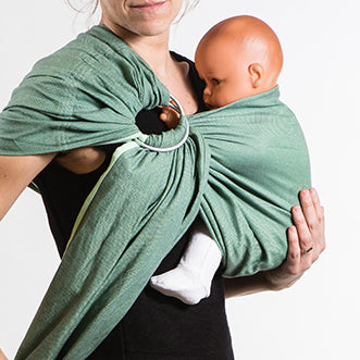 Homem a usar um sling de argolas Neobulle Vert Eucalyptus, com um bebé posicionado na frente do seu corpo, mostrando a forma como o sling envolve o corpo do bebé.