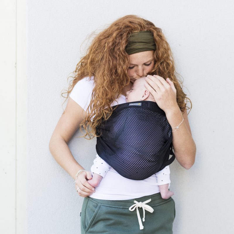 Mulher de cabelo ruivo com uma faixa na cabeça, usando um sling preto com um bebé recém-nascido encostado ao peito, de olhos fechados em gesto de carinho.