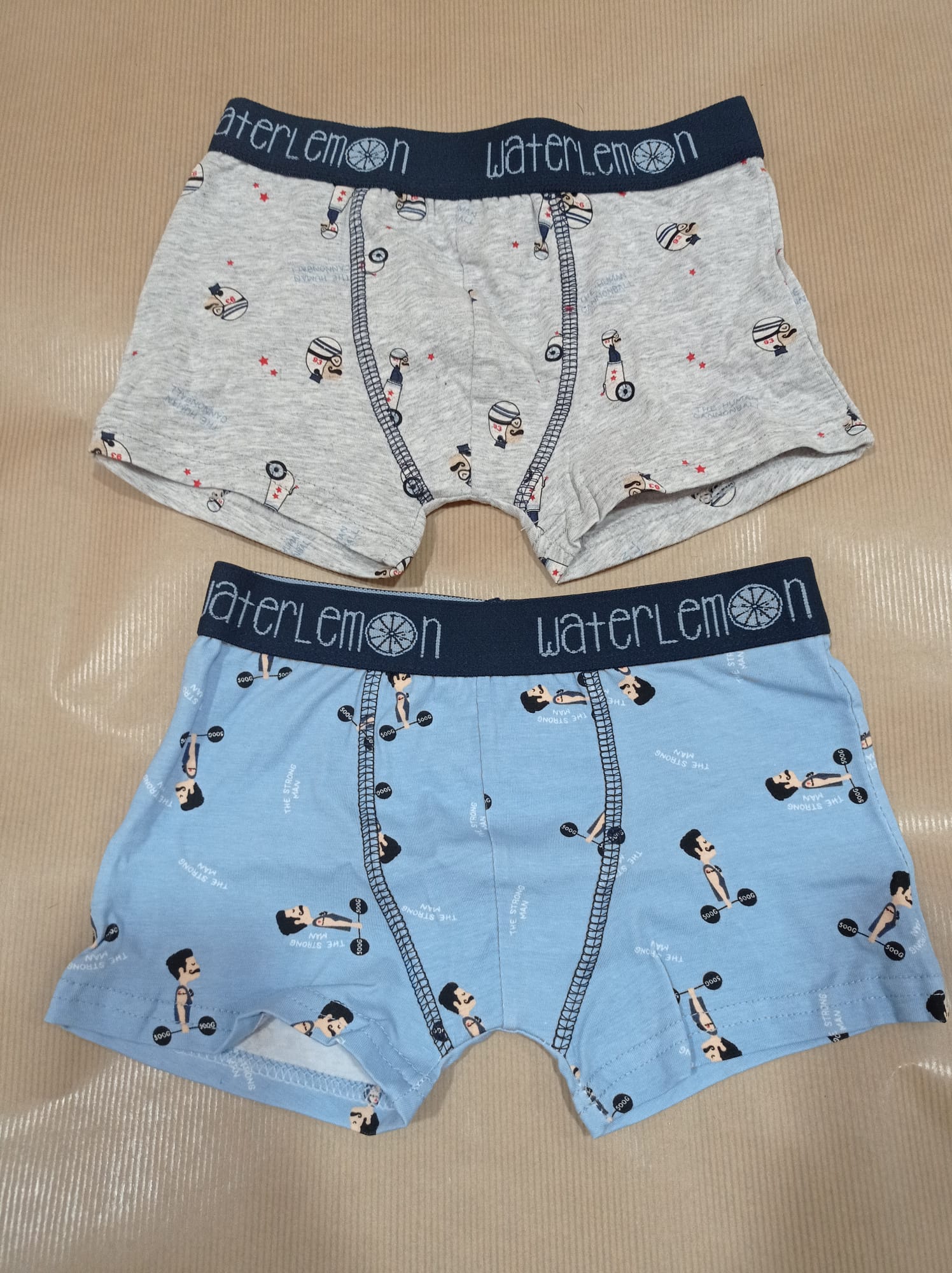 Dois boxers Waterlemon para criança, um cinzento com padrões de figuras e outro azul-claro com figuras de 'strongman'.