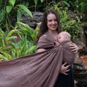 Mulher sorridente segurando um bebé num pano tecido de cor chocolate, numa posição de transporte lateral. Ambos estão rodeados por uma vegetação exuberante, o que destaca a naturalidade e conforto do produto. O pano envolve o corpo da mulher e do bebé, mostrando a sua textura macia e ajuste firme.