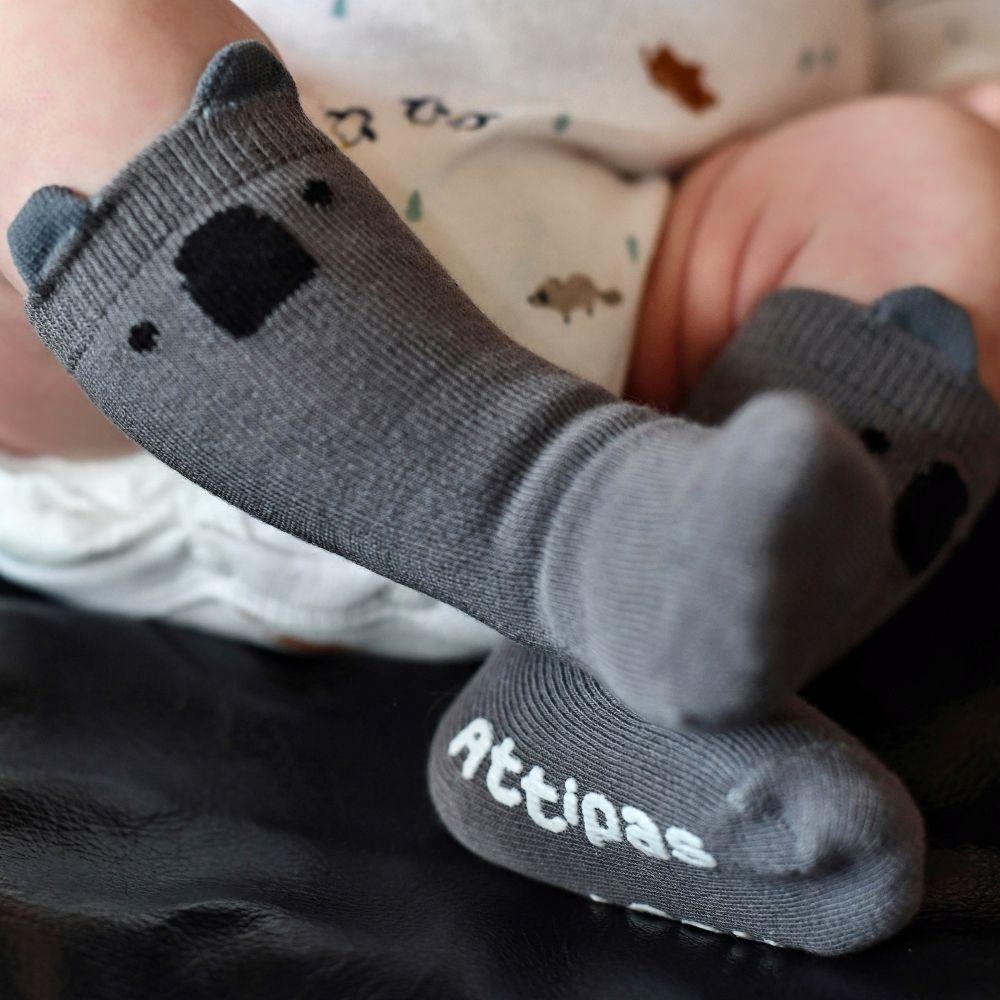 Par de meias cinzentas Attipas com detalhes de urso e sola antiderrapante com a marca Attipas no pé de um bebé.