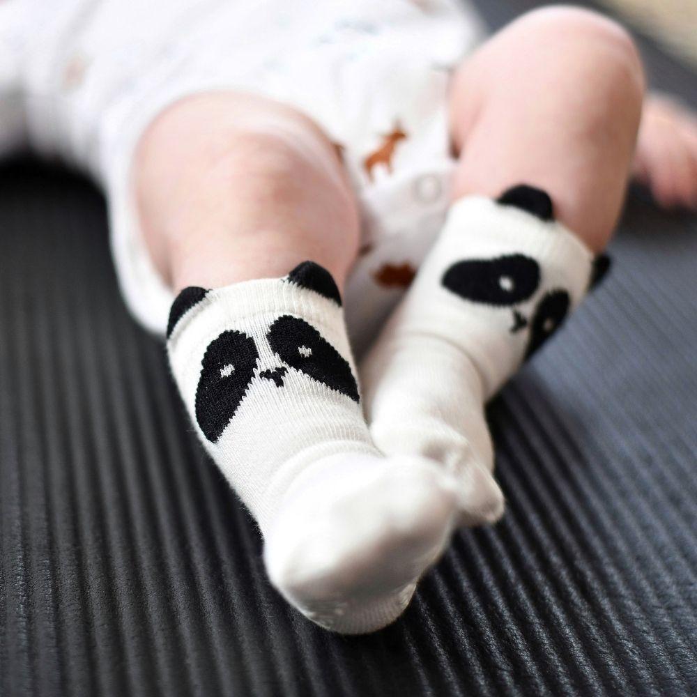Pés de um bebé a usar meias Attipas brancas com desenho de panda e orelhas pretas, em cima de um tapete escuro.