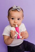 Bebé com roupa branca e laço cinzento a usar o mordedor unicórnio rosa e branco, com fundo lilás.