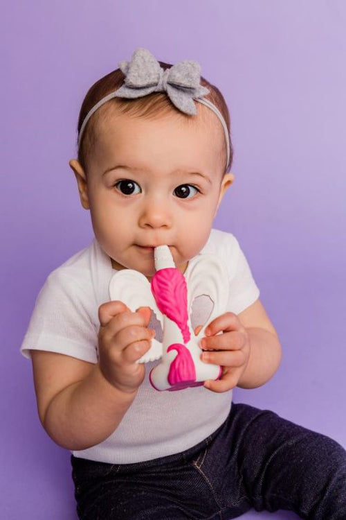 Bebé com roupa branca e laço cinzento a usar o mordedor unicórnio rosa e branco, com fundo lilás.