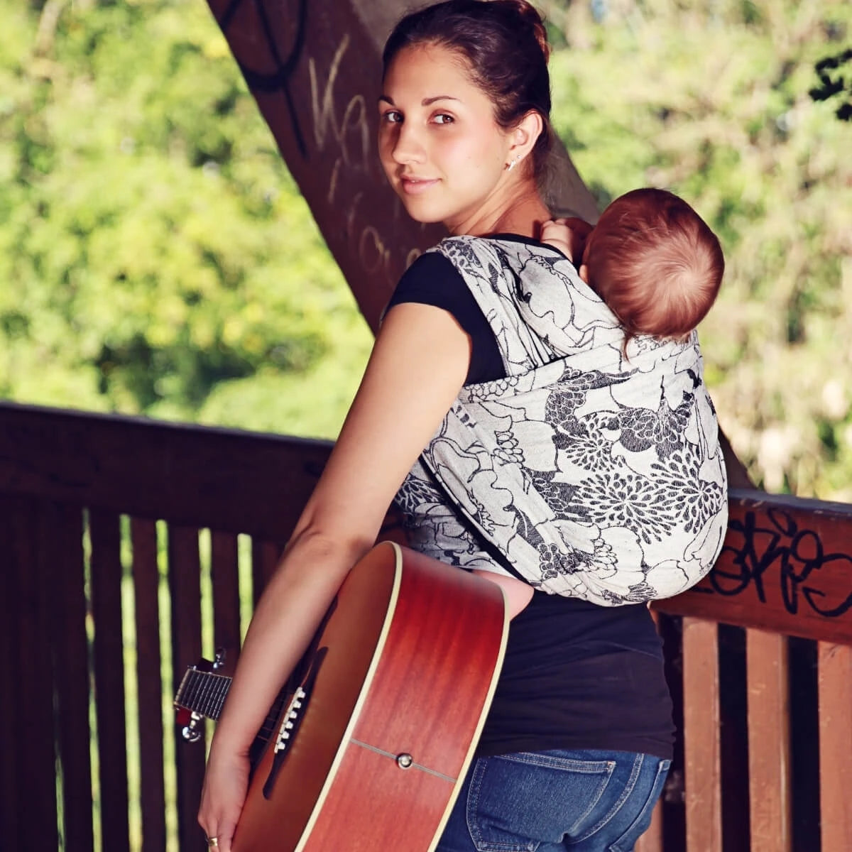 Mulher de costas com bebé no pano Hoppediz T6 Florenz, segurando uma guitarra.