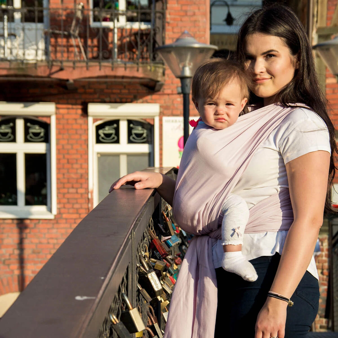 Mulher sorridente usando um pano de babywearing rosa pastel, com um bebé adormecido aconchegado contra o seu peito.