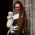 Mãe com cabelo comprido a usar o porta-bebé Mei Tai Hoppediz com um bebé pequeno.