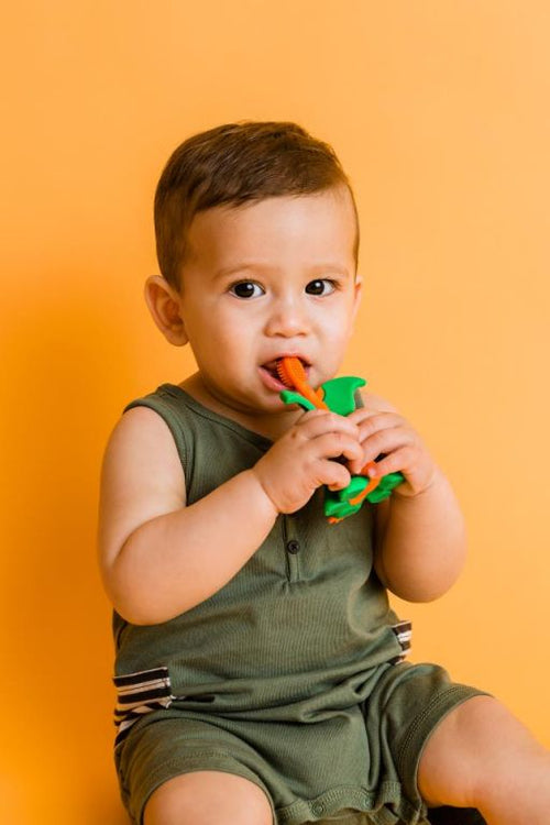 Bebé de pele escura a usar o mordedor/escova Baby Banana Dragão verde e laranja, com um fundo laranja vivo.