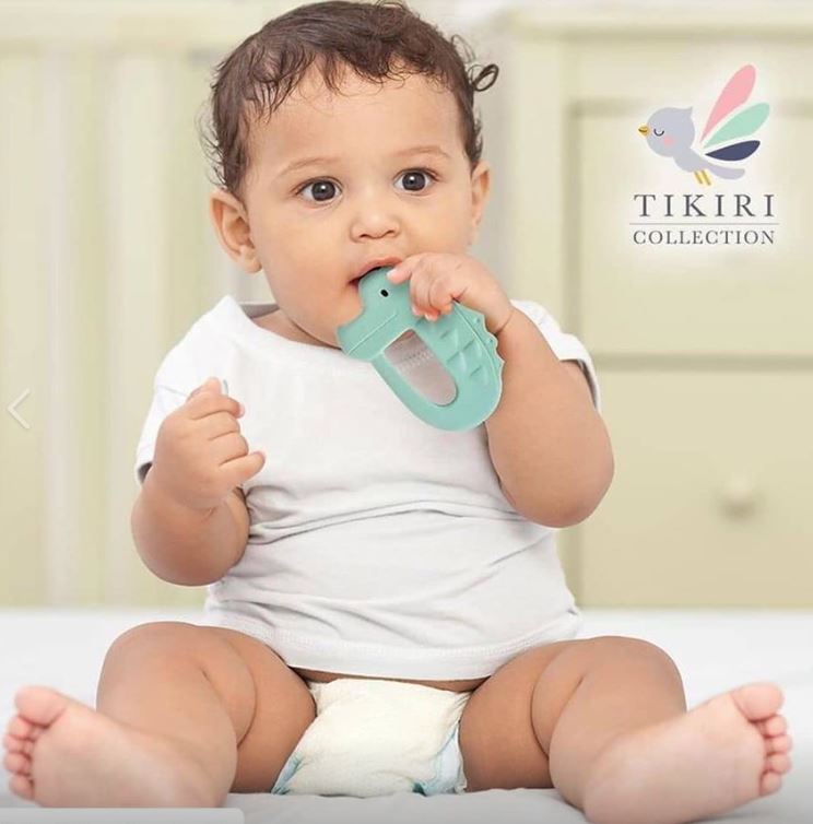 Bebé a segurar e a morder um mordedor de borracha natural verde em forma de crocodilo, com o logótipo Tikiri Collection no canto.