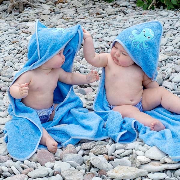 Dois bebés sentados numa praia de pedras, cada um enrolado numa toalha de banho azul com capuz de polvo.