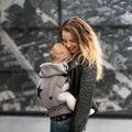 Mulher sorridente a usar o Mei Tai Hoppediz Híbrido Bondolino Plus Grey com estrela com bebé na posição frontal em ambiente urbano.