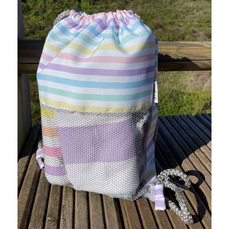 Detalhe da mochila Btbox sem areia com riscas pastel, mostrando o tecido de nylon resistente e o bolso exterior em rede, pousada sobre um banco de madeira.