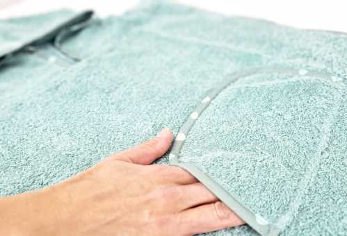 Mão a tocar a textura felpuda do poncho de algodão verde água, destacando a qualidade do tecido e a orla do bolso.