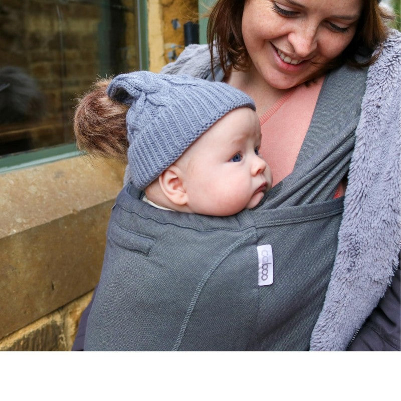 Mulher sorridente utilizando uma mochila de babywearing Caboo cinzenta clara, carregando um bebé em posição frontal.