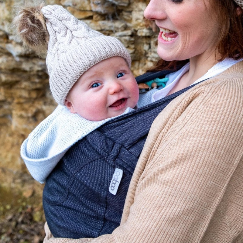 Mulher sorridente utilizando uma mochila de babywearing Caboo cinzenta clara, carregando um bebé em posição frontal, ao ar livre.