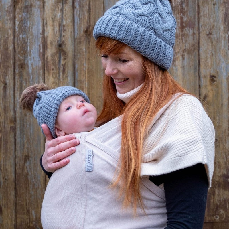 Uma mãe com cabelo ruivo a usar um porta-bebés de pano elástico, transportando o seu bebé com conforto e segurança. Ambos estão vestidos de forma casual e a mãe usa um gorro cinza.