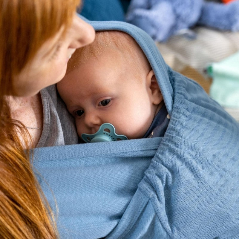 Uma mãe a usar um porta-bebés azul claro de pano elástico, com o seu bebé aconchegado e a dormir no interior.