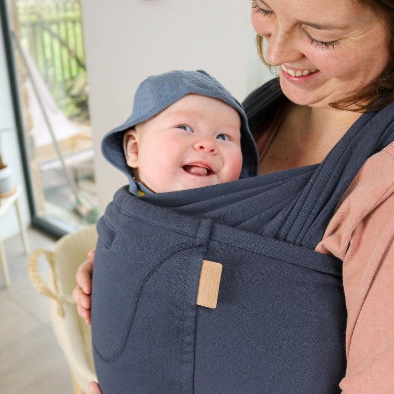 Uma mãe sorri enquanto carrega o seu bebé numa Mochila Caboo azul, proporcionando conforto e proximidade. O bebé está seguro e aconchegado com um chapéu azul.