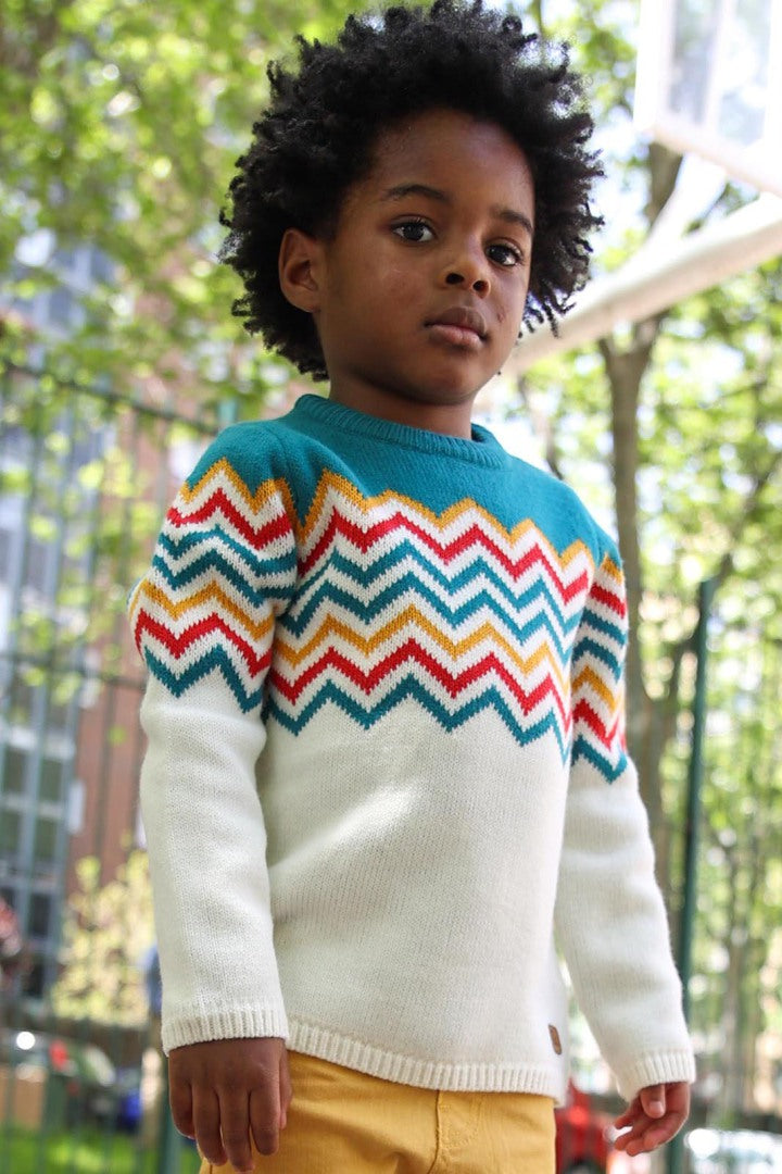 Criança a usar uma camisola de malha em tons vibrantes, com padrão zig-zag colorido. Peça confortável e quente, perfeita para os dias frios com um toque moderno.