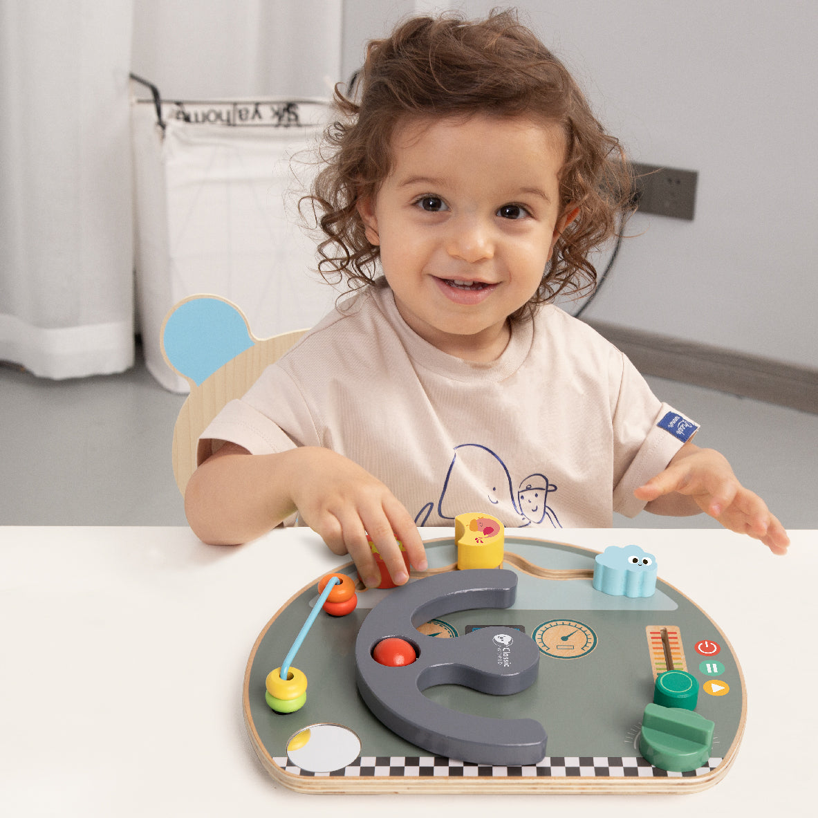 Criança sorrindo e brincando com o Classic World busy board Quadro de atividades Volante numa mesa.