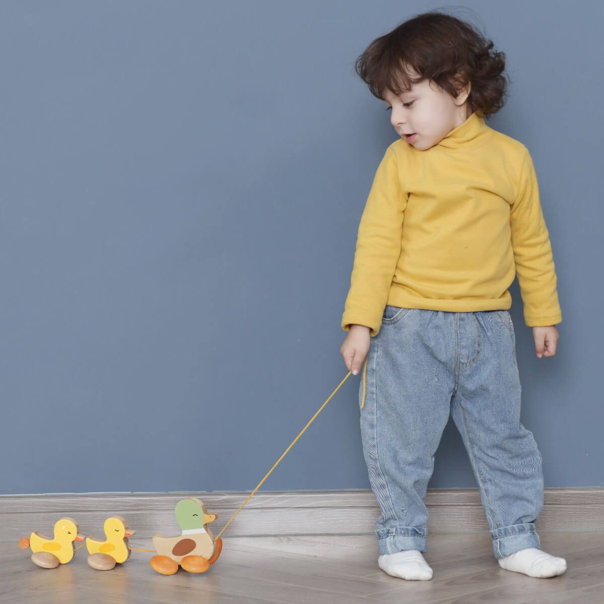 Criança pequena a puxar a família de patinhos de madeira Classic World num chão de madeira, com uma parede azul ao fundo.