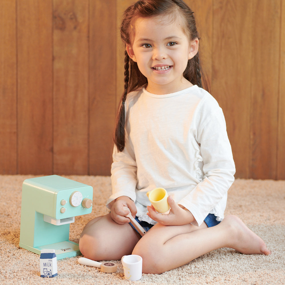Criança sorrindo ao lado da máquina de café de madeira Classic World, um brinquedo educativo e divertido.