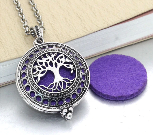 Colar difusor pessoal prateado com medalhão recortado com o padrão da Árvore da Vida ao lado de uma almofada de feltro roxa.