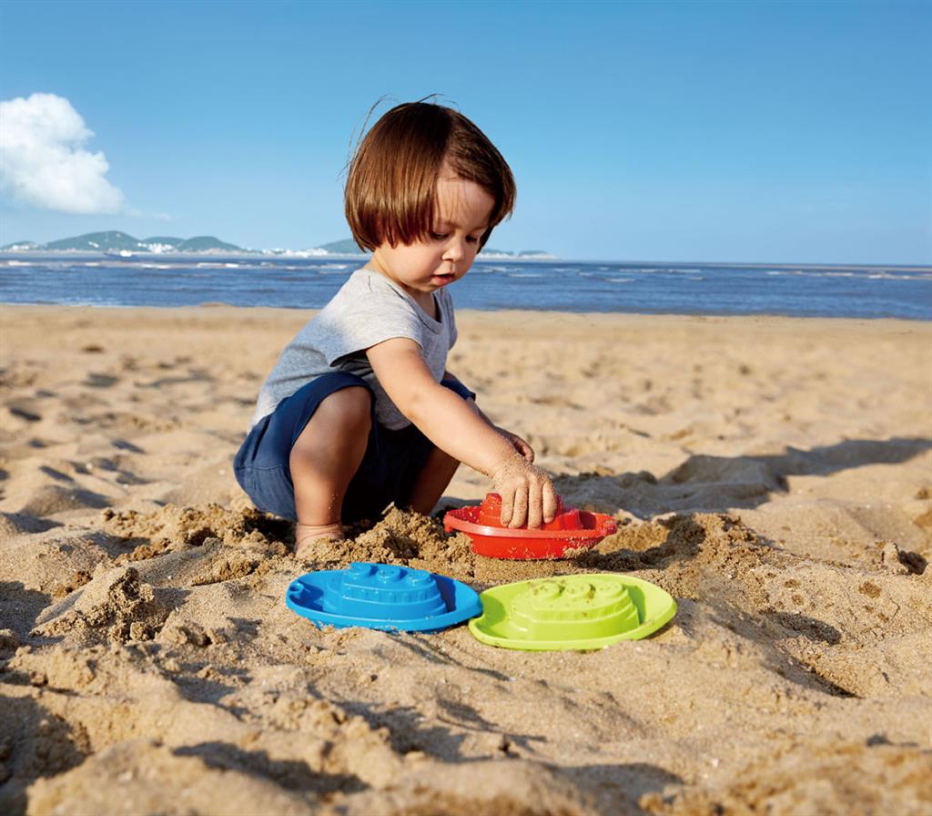Criança sentada na areia da praia a brincar com os barquinhos coloridos da Hape.