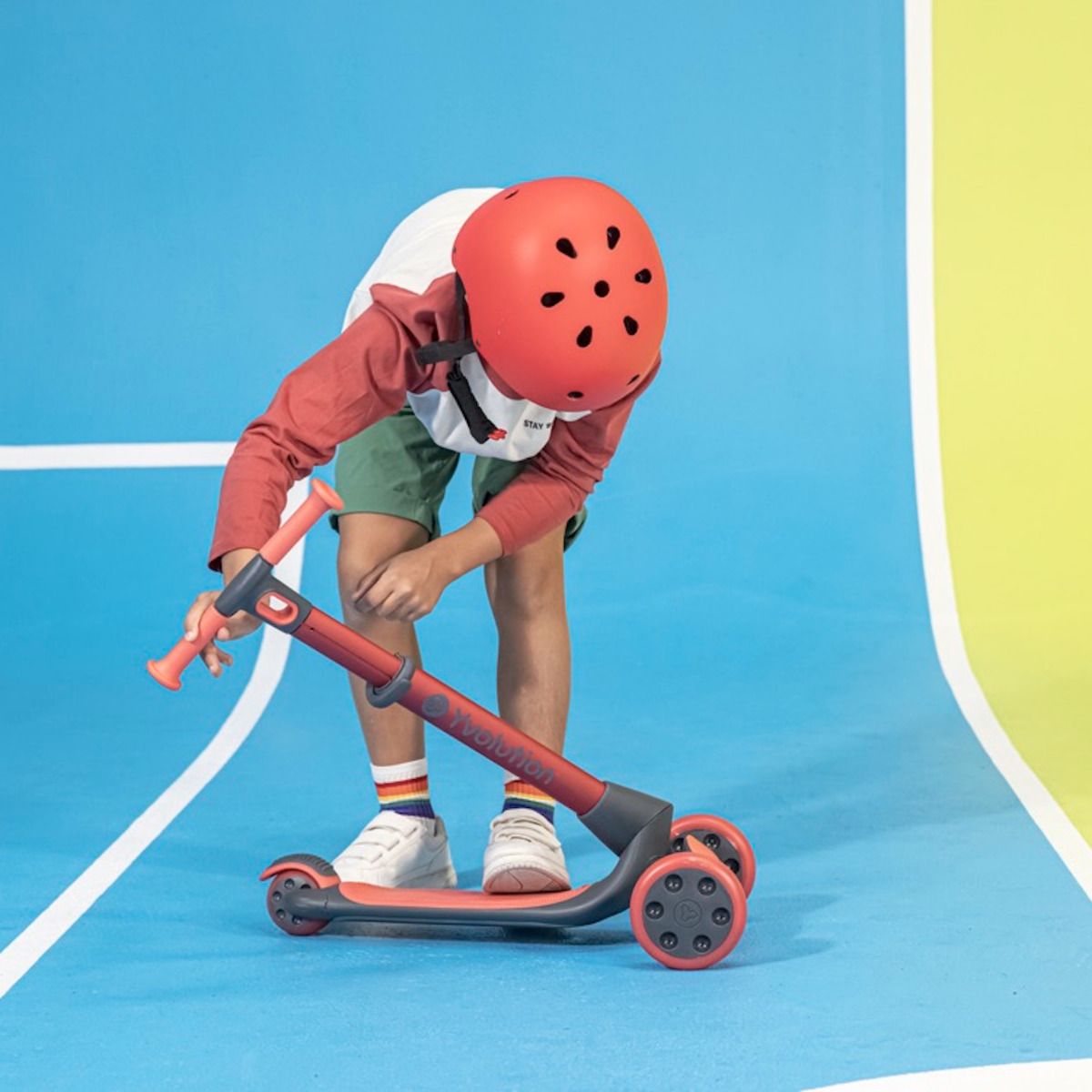 Uma criança com capacete vermelho monta o Glider Luna no modo trotinete, dobrando-o para ajustar o guiador.
