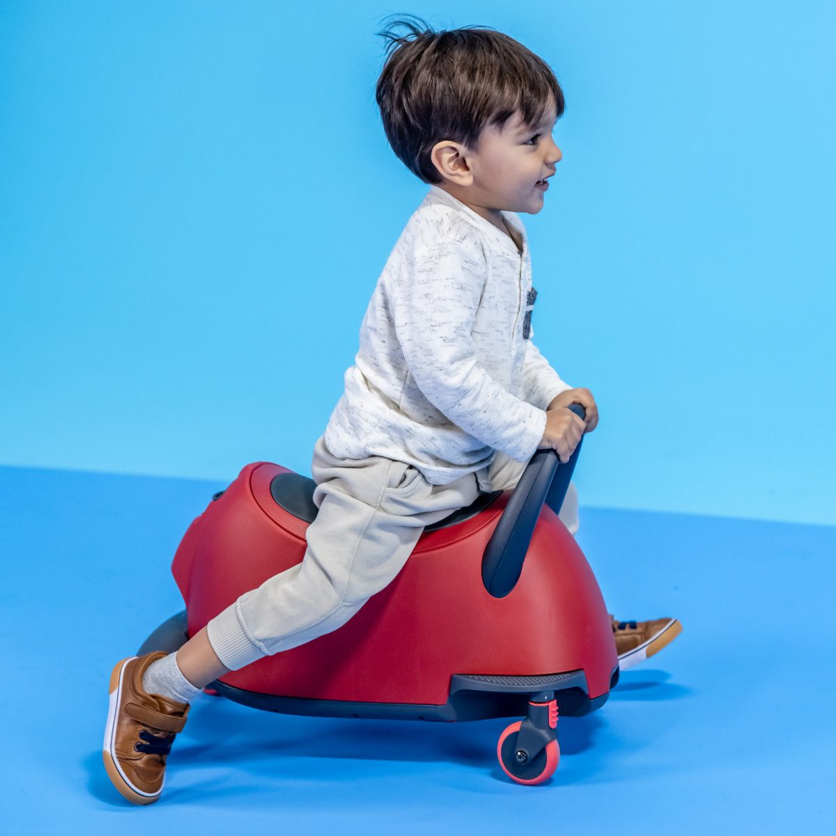 Criança pequena sorridente a andar no Glider Luna no modo "ride on", impulsionando-se com os pés, num fundo azul.