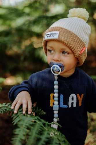 Criança a usar o gorro HELLO HOSSY com Pompom bege e castanho com pompom amarelo, na natureza.