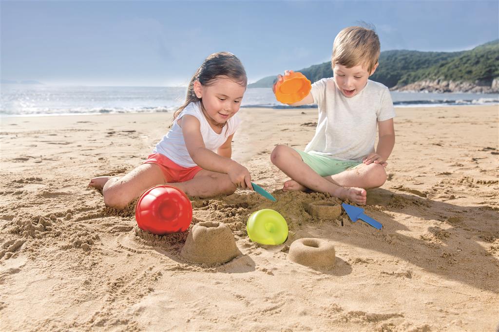 Duas crianças a brincar na areia da praia com o set de pastelaria Hape, criando bolos de areia.