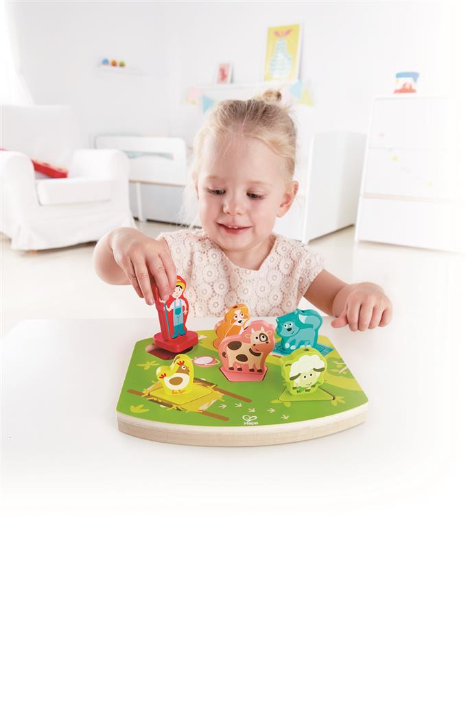 Criança sorridente a brincar com o Hape Puzzle encaixável e sonoro A Quinta, colocando a peça do agricultor na base.