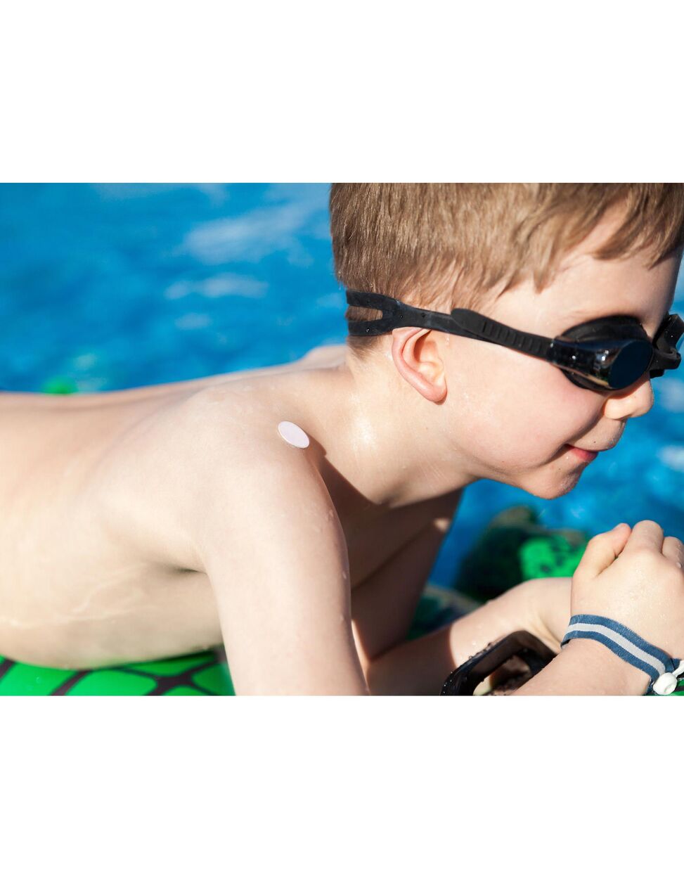Criança com adesivo Everyday Baby SunHero no ombro, indicador de proteção solar. Piscina ao fundo.