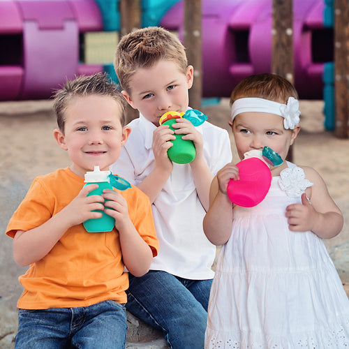 Três crianças a beber de garrafas Squeasy coloridas num parque infantil.