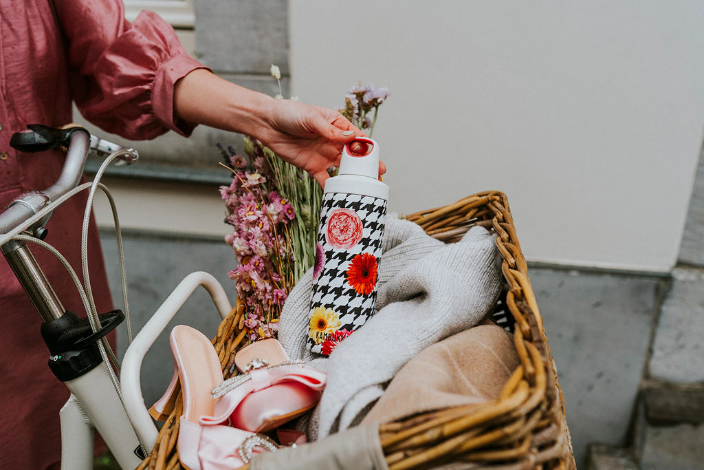Garrafa térmica Kambukka Reno 500ml com padrão floral sendo colocada num cesto de bicicleta com flores e sapatos, mostrando o uso diário.