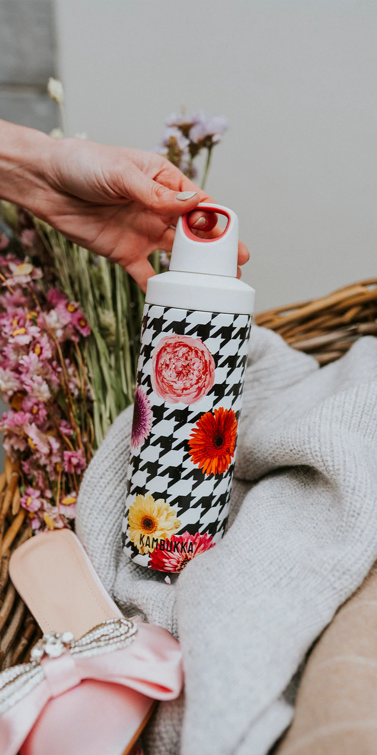Garrafa térmica Kambukka Reno 500ml com padrão xadrez preto e branco e flores coloridas, sendo segurada por uma mão sobre um cesto com têxteis.