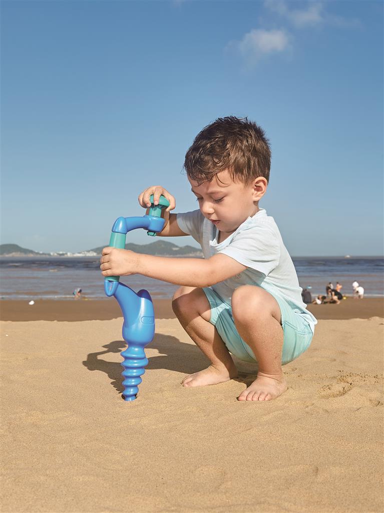 Criança (mãos visíveis) a usar o furador Hape na areia, a girar a pega amarela para estimular a motricidade fina e o jogo imaginativo.