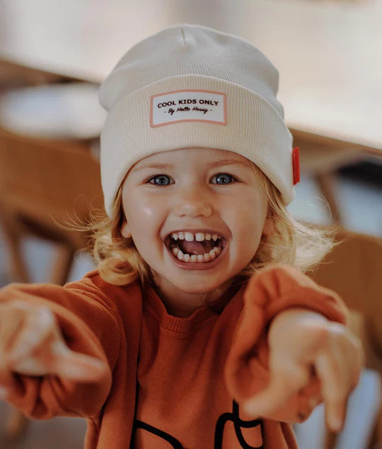 Criança feliz a rir e a apontar, a usar gorro creme Hello Hossy. Mostra gorro criança em uso.