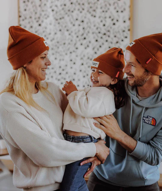 Família (pai, mãe, filha) a interagir feliz, todos com gorro laranja Hello Hossy. Mostra gorro criança em contexto familiar.