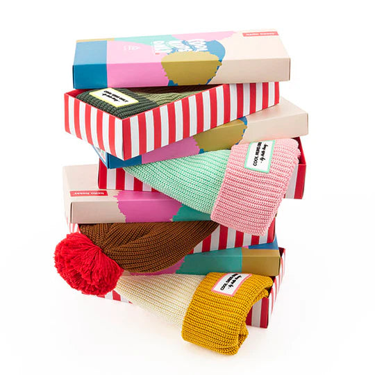 Pilha de caixas com gorros HELLO HOSSY com Pompom em várias cores e padrões.