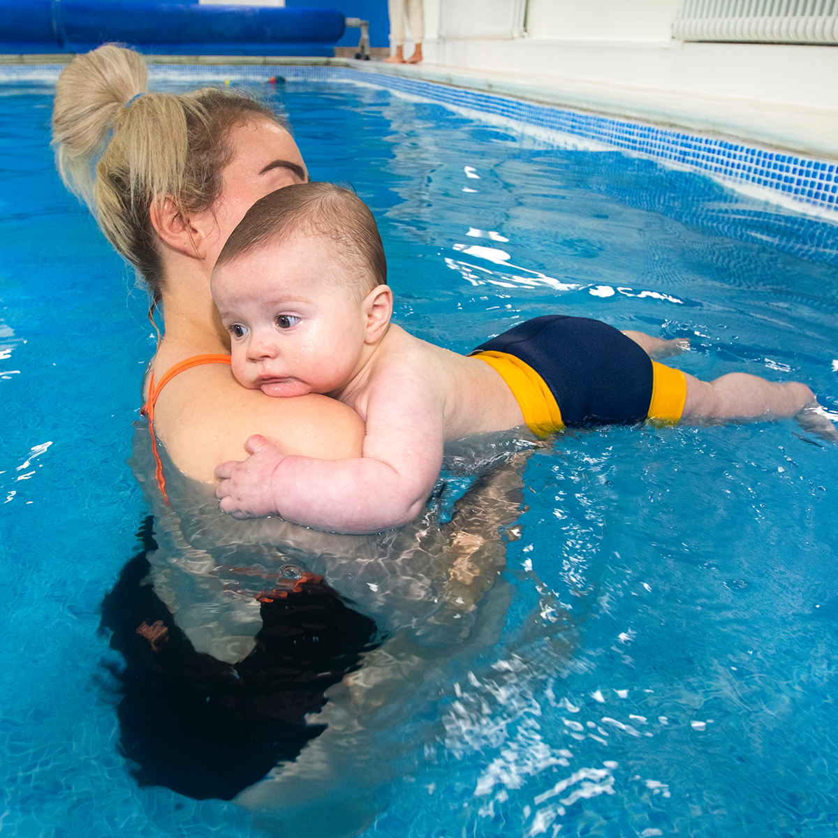 Bebé a flutuar na piscina, apoiado no ombro de um adulto, usando a fralda de natação Splash About Happy Nappy DUO Navy/Yellow.