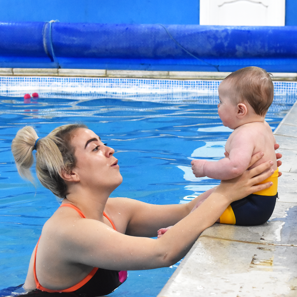 Bebé com a fralda de natação Splash About Happy Nappy DUO Navy/Yellow sentado na borda da piscina, interagindo com um adulto.