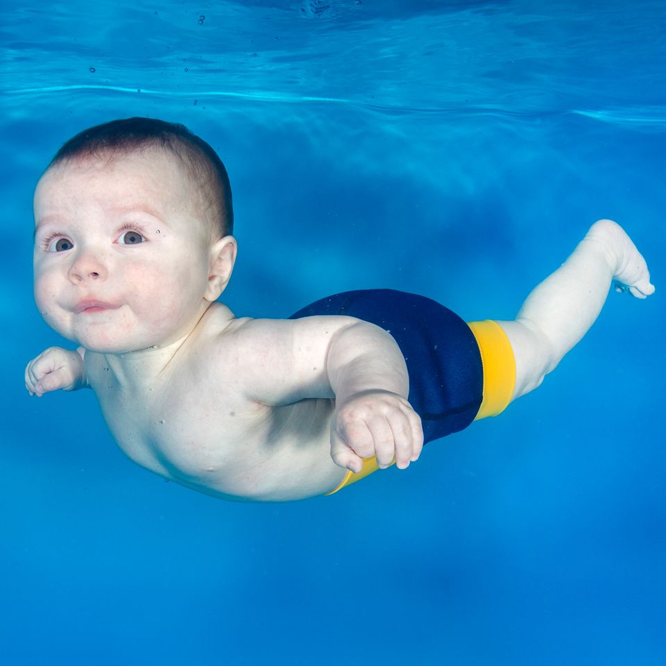 Bebé com a fralda de natação Splash About Happy Nappy DUO Navy/Yellow a nadar debaixo de água, com bolhas visíveis.