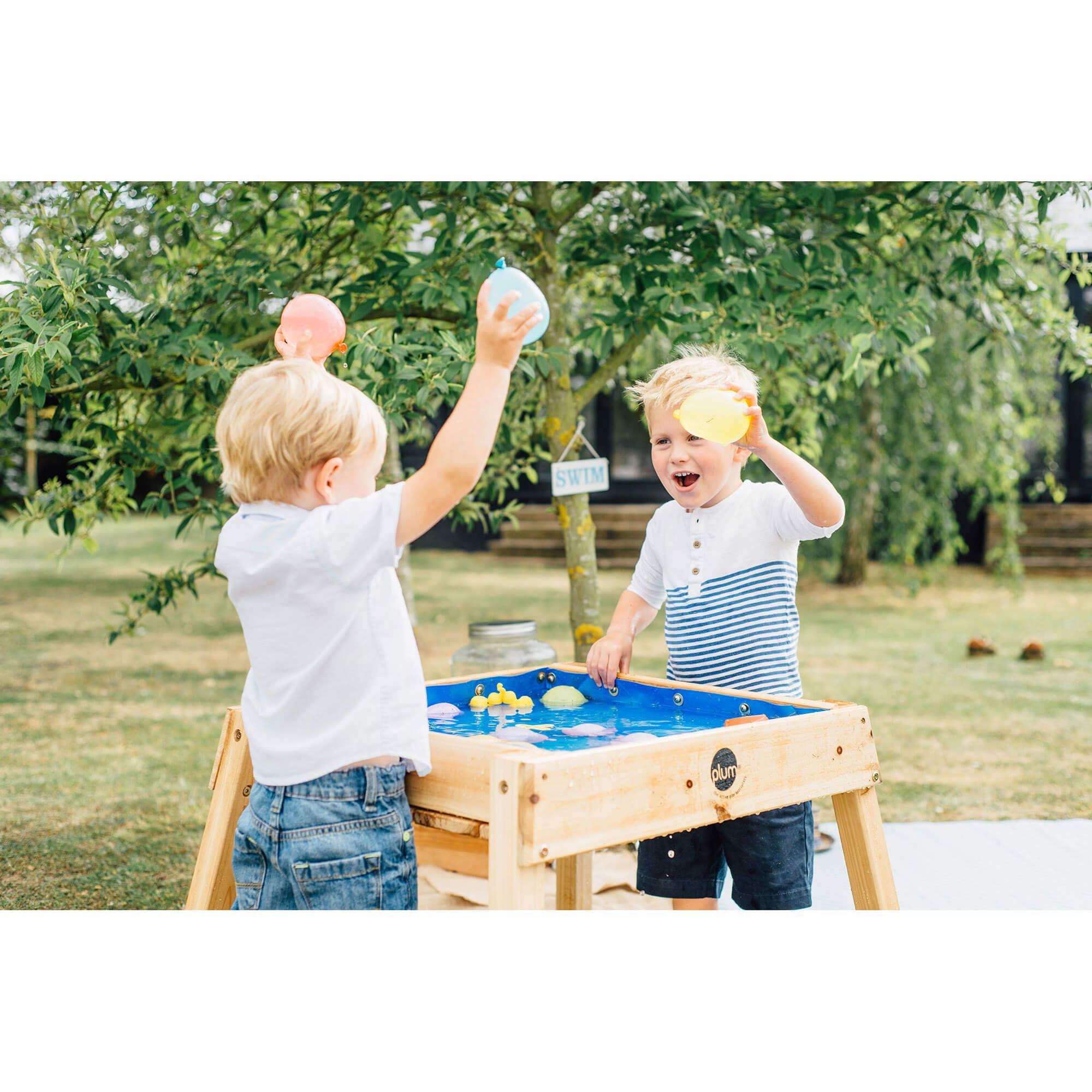 Duas crianças felizes a brincar com balões de água na mesa Plum multiusos para ar livre, num dia ensolarado no jardim.