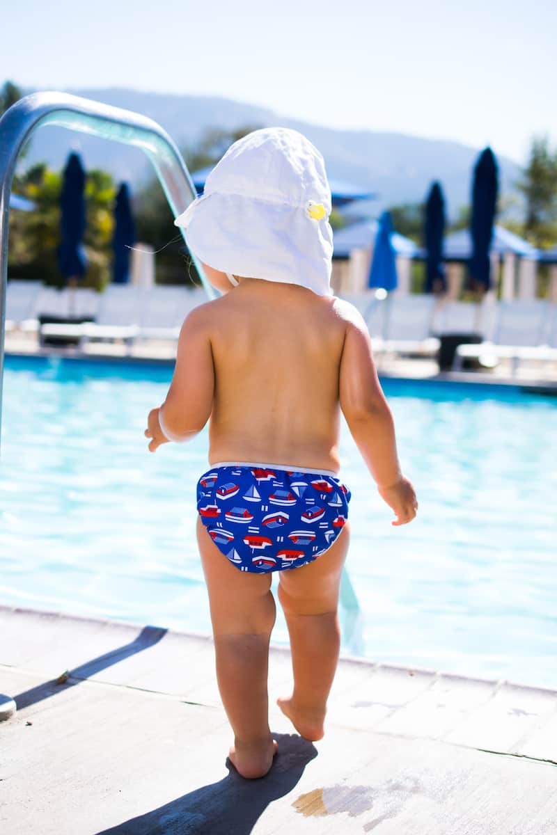 Bebé a caminhar junto à piscina com fralda de banho ImseVimse e chapéu de proteção.
